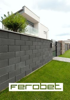 a large gray brick wall in front of a house with green grass and bushes on the side