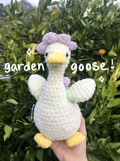 a crocheted white duck with a purple hat on it's head is held in front of some bushes