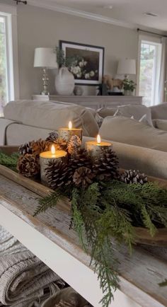 candles are lit on a tray with pine cones and greenery in front of a couch