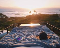 a bed with a hat on top of it next to the ocean at sunset or sunrise