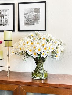 a vase filled with white daisies sitting on top of a wooden table next to two framed pictures