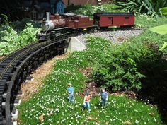 three miniature people are standing in the grass near a toy train track with flowers on it