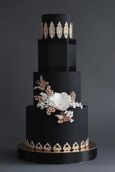 a three tiered black cake with white flowers and leaves on the top, in front of a gray background