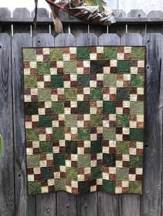 a green and brown quilt hanging on a wooden fence next to a potted plant