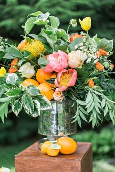 a vase filled with flowers and oranges on top of a wooden block next to trees