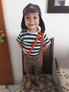 a young boy wearing a hat and suspenders standing on top of a wooden chair