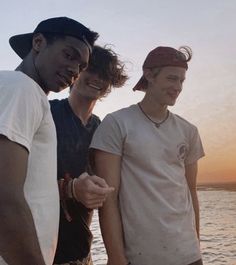 three young men standing next to each other near the ocean at sunset, one wearing a hat