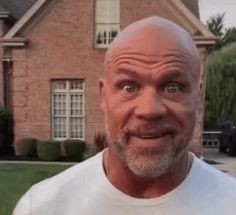 a bald man in a white t - shirt standing in front of a brick house