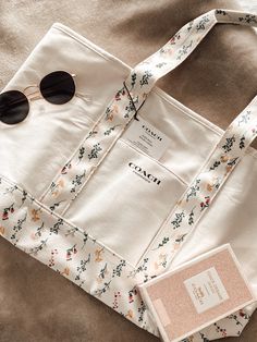 a white tote bag sitting on top of a bed next to a pair of sunglasses