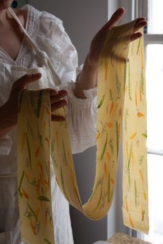 a woman is holding up a yellow scarf with green and orange designs on it while standing in front of a window