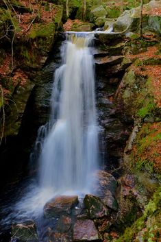 a small waterfall in the middle of a forest with rocks and trees around it on a fall day