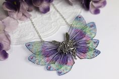 a purple and green butterfly necklace sitting on top of a white doily next to purple flowers