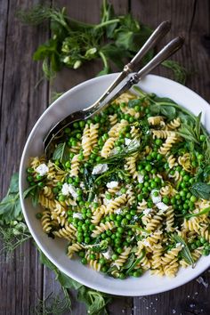 a bowl filled with pasta and peas on top of a wooden table next to two spoons