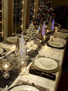 a table set for christmas dinner with silver and white plates, candles and trees in the background