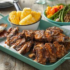 a tray filled with meat and vegetables on top of a wooden table next to a bowl of corn