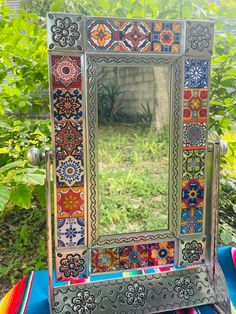 a colorful mirror sitting on top of a table next to a tree and bushes in the background