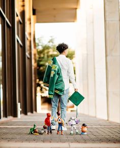 a young boy is walking down the sidewalk with his toy figures and green jacket on