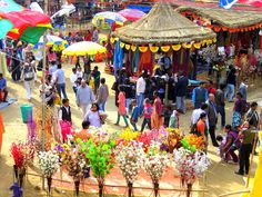 a crowd of people walking around a market with umbrellas and flowers in the foreground