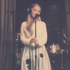 a woman in a white dress holding a microphone and standing next to a drum set