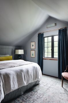 a bed sitting under a window in a bedroom next to a chair and table with a lamp on it