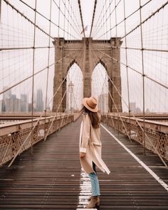 a woman walking across a bridge in front of the brooklyn bridge with her hat on