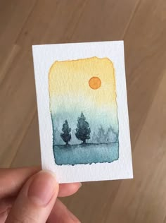 a hand holding up a small card with trees on it and the sun in the background