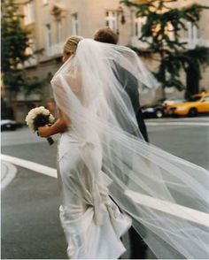 a bride and groom walking down the street in their wedding gowns, veils blowing in the wind