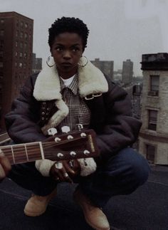 a man sitting on top of a roof with an acoustic guitar in his hand and buildings in the background