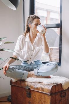 a woman sitting on top of a wooden trunk drinking from a cup while holding a coffee mug