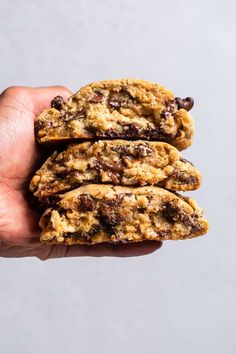 a hand holding three chocolate chip cookies on top of each other in front of a gray background