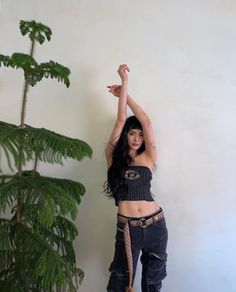 a woman in black top and jeans standing next to a potted plant with her arms up