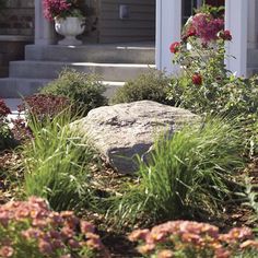 a rock sitting in front of a house with flowers around it on the side walk