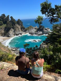 two people sitting on the edge of a cliff looking out at an ocean and beach