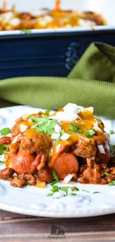 a white plate topped with chili and cheese covered hot dog casserole next to a green napkin