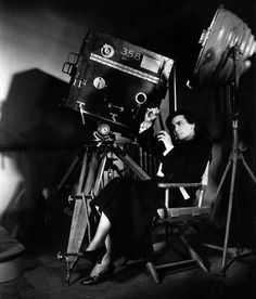 a black and white photo of a woman sitting in a chair next to a camera