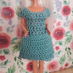 a doll wearing a crocheted dress and white hair standing in front of a floral wallpaper