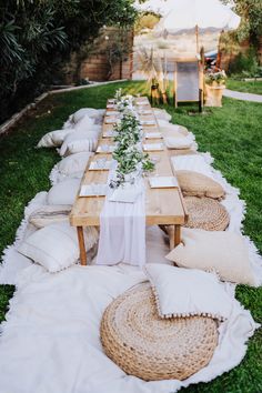 a long table set up with white linens, pillows and greenery on the grass