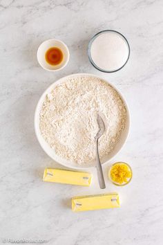 ingredients to make an orange cake laid out on a white marble counter top, including flour, butter, and eggs