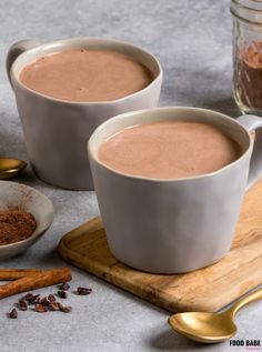 two mugs of hot chocolate on a cutting board next to cinnamon sticks and spices