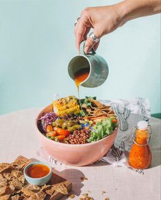 a person pouring dressing into a bowl filled with vegetables and crackers on a table