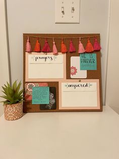 a bulletin board with notes and tassels on it next to a potted plant