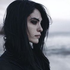 a woman with long black hair and dark makeup looks off into the distance while standing in front of water