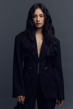 a woman with long hair wearing a black jacket and pants, standing in front of a gray background