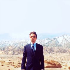 a man in a suit and tie standing on top of a desert area with mountains in the background