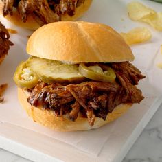 a pulled pork sandwich with pickles and onions on a cutting board next to chips