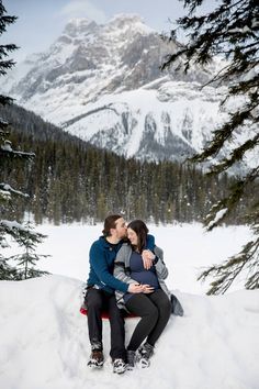 a man and woman sitting on top of a snow covered hill next to pine trees