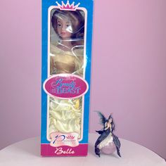 a doll in a box next to a fairy figurine on a white table