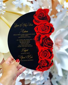 a woman holding up a black and red wedding card with roses on the front, in front of white flowers