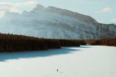 a person walking across snow covered ground next to tall pine trees and mountain in the background