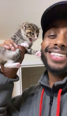 a man holding a kitten up to his face while wearing a hat and smiling at the camera